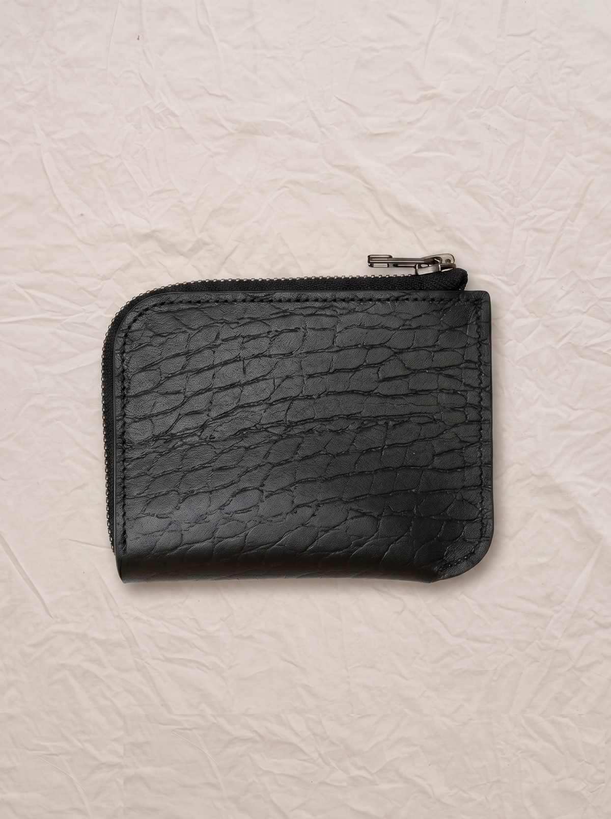 A small black zippered pouch with a crocodile-embossed texture, photographed flat on a wrinkled off-white paper background.